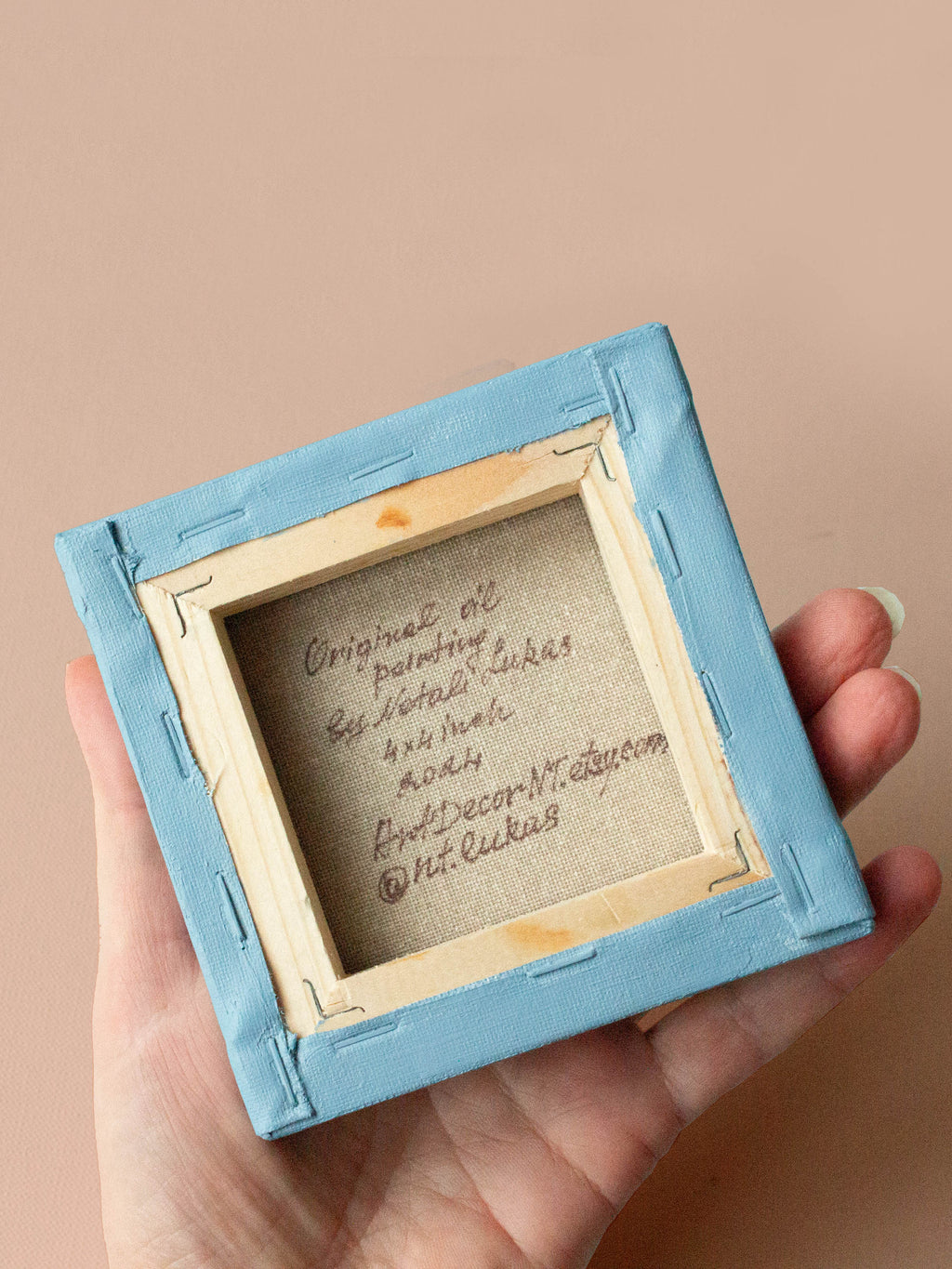 Hand holding a small blue-framed artwork with visible text on a beige background
