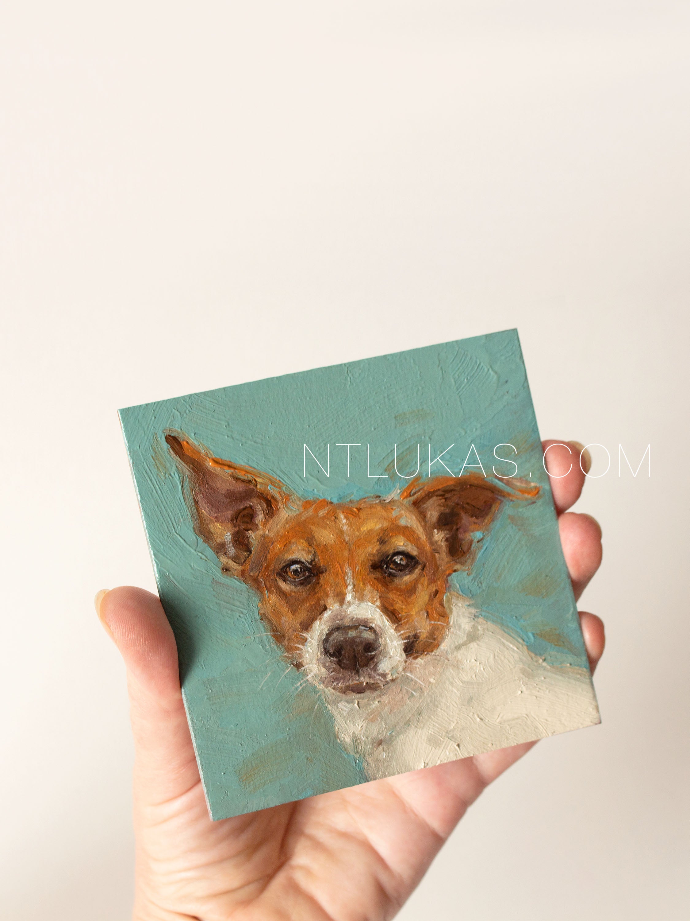 Hand holding a square painting of a dog's face with a teal background.